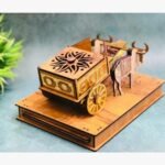 Intricately carved wooden bullock cart miniature sculpture featuring gold accents and traditional Indian craftsmanship on a decorative pedestal.