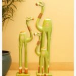 A set of three hand-carved wooden camel sculptures in varying sizes, painted in a soft matte green with jute neck accents and floral motifs.