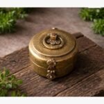 A circular hand-engraved antique brass spice or jewelry box featuring intricate traditional patterns and a hinged clasp on a rustic wood surface.