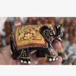 A miniature wooden elephant figurine intricately hand-painted with traditional gold, red, and black floral patterns, held in a palm.