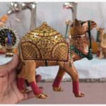 A close-up of a handcrafted wooden camel figurine, featuring detailed gold-leaf style floral painting and vibrant red leg accents.
