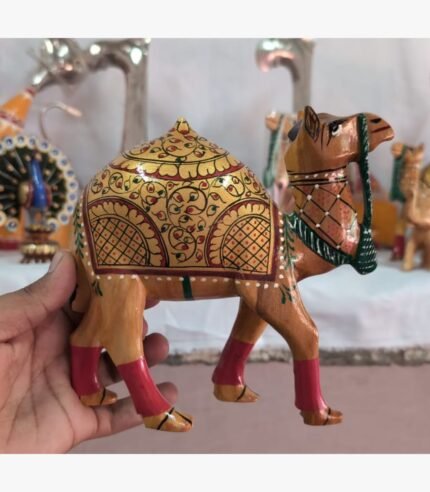 A close-up of a handcrafted wooden camel figurine, featuring detailed gold-leaf style floral painting and vibrant red leg accents.
