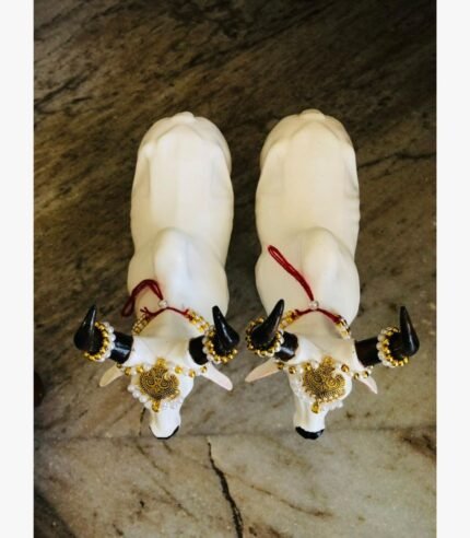A top-down view of two white marble-finish Nandi bull sculptures adorned with traditional golden jewelry, pearls, and black horns.