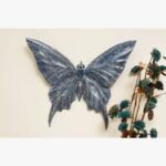 A hand-carved wooden butterfly wall sculpture featuring a distressed indigo blue finish and intricate wing textures, displayed on a cream wall.