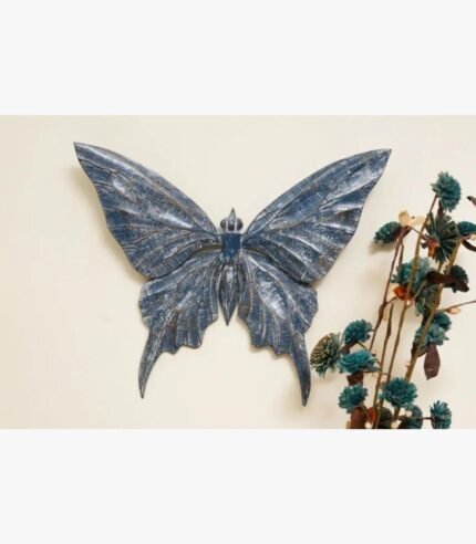 A hand-carved wooden butterfly wall sculpture featuring a distressed indigo blue finish and intricate wing textures, displayed on a cream wall.
