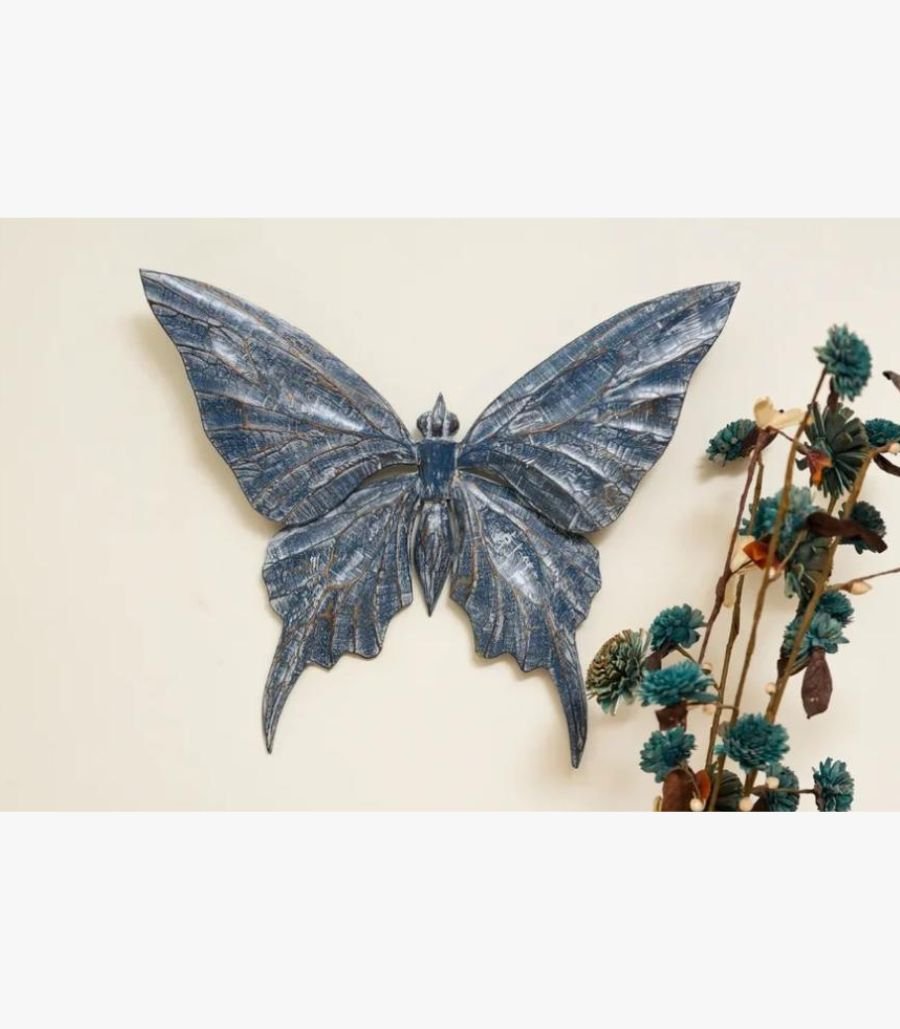 Indigo Azure Butterfly Wooden Wall Art A hand-carved wooden butterfly wall sculpture featuring a distressed indigo blue finish and intricate wing textures, displayed on a cream wall.