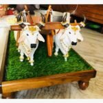 Handcrafted white Nandi bull pair with a wooden plough on a green turf base, featuring gold jewelry and pearl embellishments.