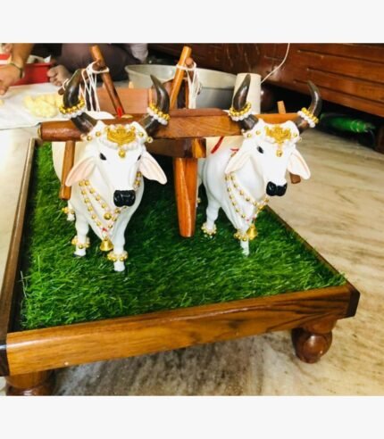 Handcrafted white Nandi bull pair with a wooden plough on a green turf base, featuring gold jewelry and pearl embellishments.