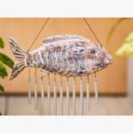 A hand-carved wooden fish wind chime with a distressed white and brown finish featuring hanging metallic chimes for coastal home decor.