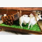 A side-profile view of a premium handcrafted wooden bullock cart pulled by two adorned white Nandi bulls on a green turf pedestal base.