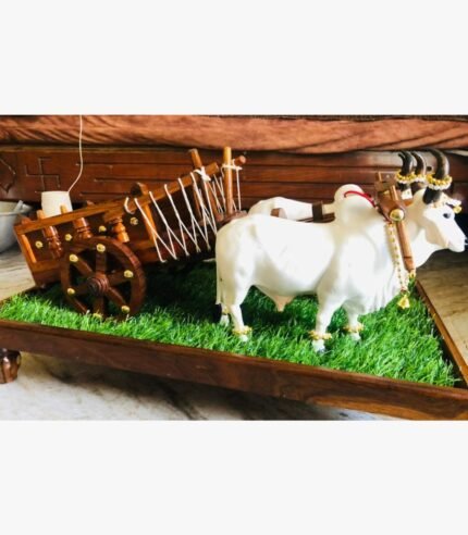 A side-profile view of a premium handcrafted wooden bullock cart pulled by two adorned white Nandi bulls on a green turf pedestal base.