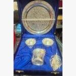 A premium 5-piece Ornate German Silver Pooja Thali set featuring an etched ritual plate, bowls, a pedestal diya, and a feather-shaped applicator in a velvet presentation box.