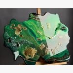 Luxury custom-shaped green geode epoxy resin wall clock titled Emerald Zodiac by Vina Bhavsar featuring gold Scorpio and Taurus astrological signs, golden Roman numerals, and textured emerald crushed crystal clusters.