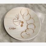 Handcrafted circular decorative plate titled Caffeine Rings by Vina Bhavsar featuring a minimalist cream background with a stylized coffee cup sketch, coffee beans, and overlapping brown coffee cup stain rings.