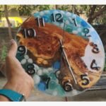 Handcrafted circular resin river wall clock titled Aqua Pebble by Vina Bhavsar featuring a live edge burl wood slab embedded with smooth white pebbles and blue glass stones.