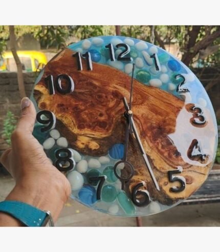 Handcrafted circular resin river wall clock titled Aqua Pebble by Vina Bhavsar featuring a live edge burl wood slab embedded with smooth white pebbles and blue glass stones.