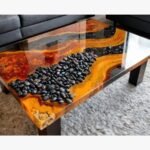 Luxury live edge burl wood coffee table titled Lava Flow by Vina Bhavsar featuring a black epoxy resin river channel filled with smooth black river stones under a high-gloss finish.