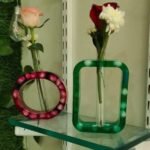 A pair of handcrafted minimalist epoxy resin bud vases titled Flora Loop by Vina Bhavsar, featuring one circular pink frame and one rectangular green frame embedding smooth stones with glass test tube inserts holding fresh flowers.