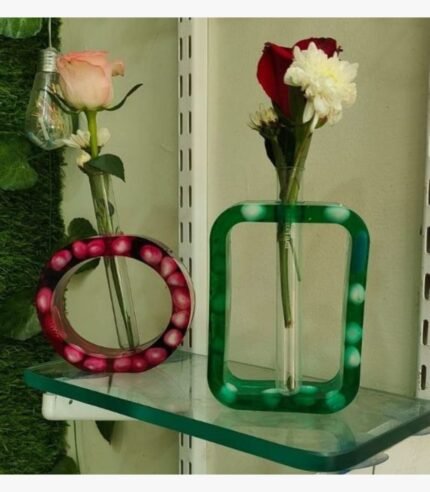 A pair of handcrafted minimalist epoxy resin bud vases titled Flora Loop by Vina Bhavsar, featuring one circular pink frame and one rectangular green frame embedding smooth stones with glass test tube inserts holding fresh flowers.