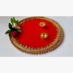 Handcrafted circular red decorative pooja thali platter titled Crimson Festive Elegance by Khushboo Gohel featuring ornate gold gota patti lace, golden bead trim, two brass diyas, and a faux rose accent.