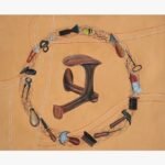 Original contemporary surrealist watercolor painting titled The Cobbler's Orbit by Vala Yash showcasing an antique iron shoe last enclosed within a circular loop of traditional leather crafting tools against a warm tan background with stitched line patterns.