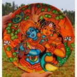 Hand-painted decorative wall plate titled Eternal Radha Krishna by Akshaya Sivadas featuring a traditional Kerala mural portrait of a blue Lord Krishna playing the flute beside Radha under a sacred tree.