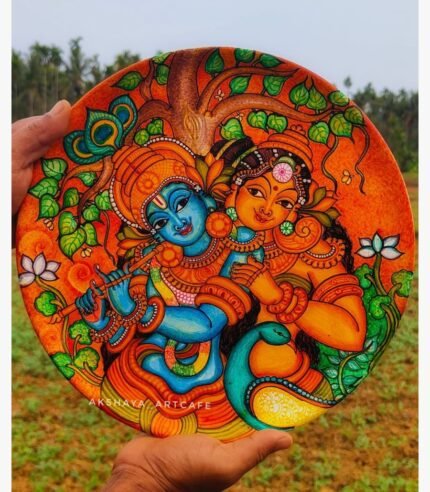 Hand-painted decorative wall plate titled Eternal Radha Krishna by Akshaya Sivadas featuring a traditional Kerala mural portrait of a blue Lord Krishna playing the flute beside Radha under a sacred tree.