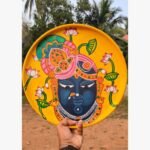 Hand-painted decorative terracotta plate titled Shrinathji Lotus Grace by Akshaya Sivadas featuring a dark-complexioned portrait of Lord Shrinathji decorated with pink lotuses and an ornate headdress on a bright yellow background.