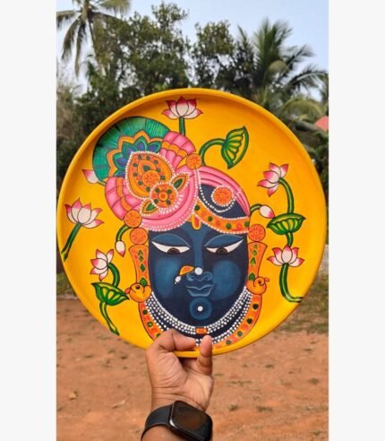 Hand-painted decorative terracotta plate titled Shrinathji Lotus Grace by Akshaya Sivadas featuring a dark-complexioned portrait of Lord Shrinathji decorated with pink lotuses and an ornate headdress on a bright yellow background.