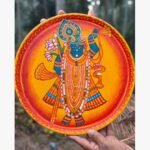 Hand-painted decorative terracotta plate titled Shrinathji Divine Radiance by Akshaya Sivadas featuring a full-body standing portrait of Lord Shrinathji with a peacock feather and lotus blossoms against a glowing orange and yellow backdrop.