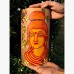 Hand-painted bamboo container titled Serene Buddha by Akshaya Sivadas featuring a detailed warm yellow and orange portrait of Lord Buddha in a traditional mural style flanked by small yellow flowers and green leaves.