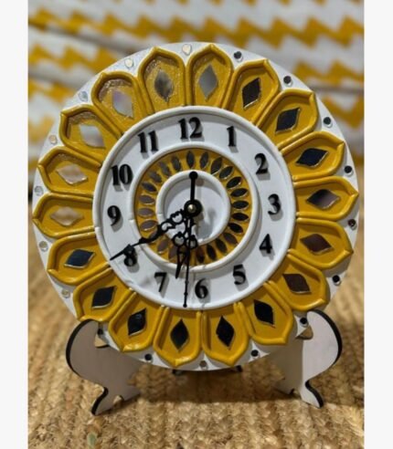Handcrafted sunflower-inspired round Lippan art table clock with mustard yellow clay relief work, white dial, and glass mirror inlays on a wooden display stand by Rucha Desai.